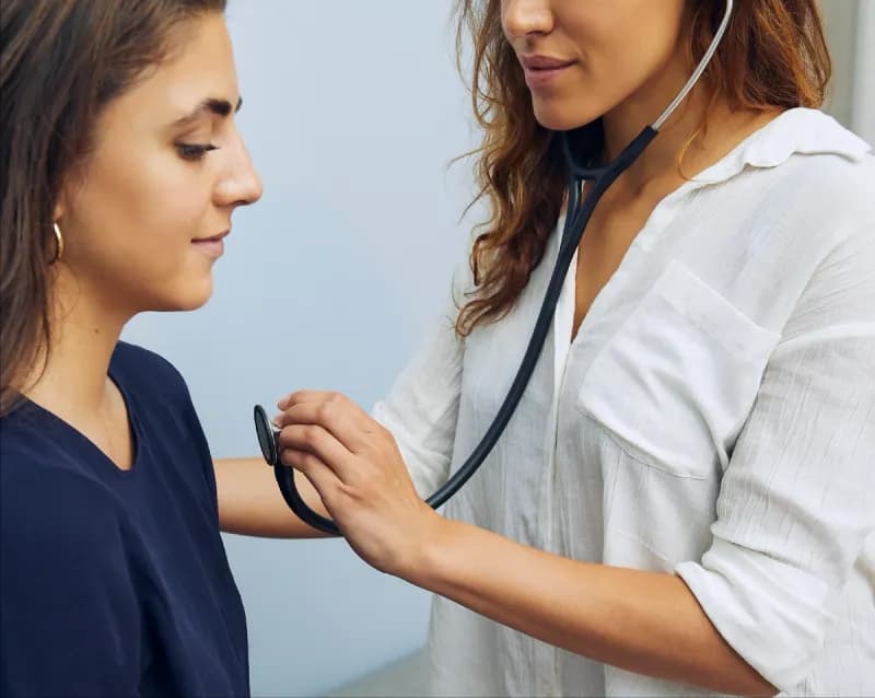 A female doctor in a white blouse uses a stethoscope to listen to the heartbeat of a calm female patient during a medical check-up.