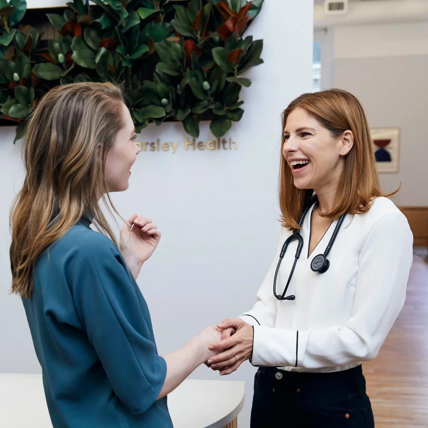A smiling female doctor wearing a stethoscope warmly greets a female patient inside a modern medical clinic with a leafy wall decoration and the name 'Parsley Health' visible in the background.